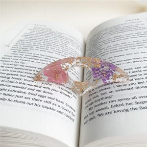 A resin book page holder featuring pink and purple flowers with gold flecks, placed between the pages of an open book. The holder delicately holds the pages in place, adding a beautiful and elegant touch to the reading experience