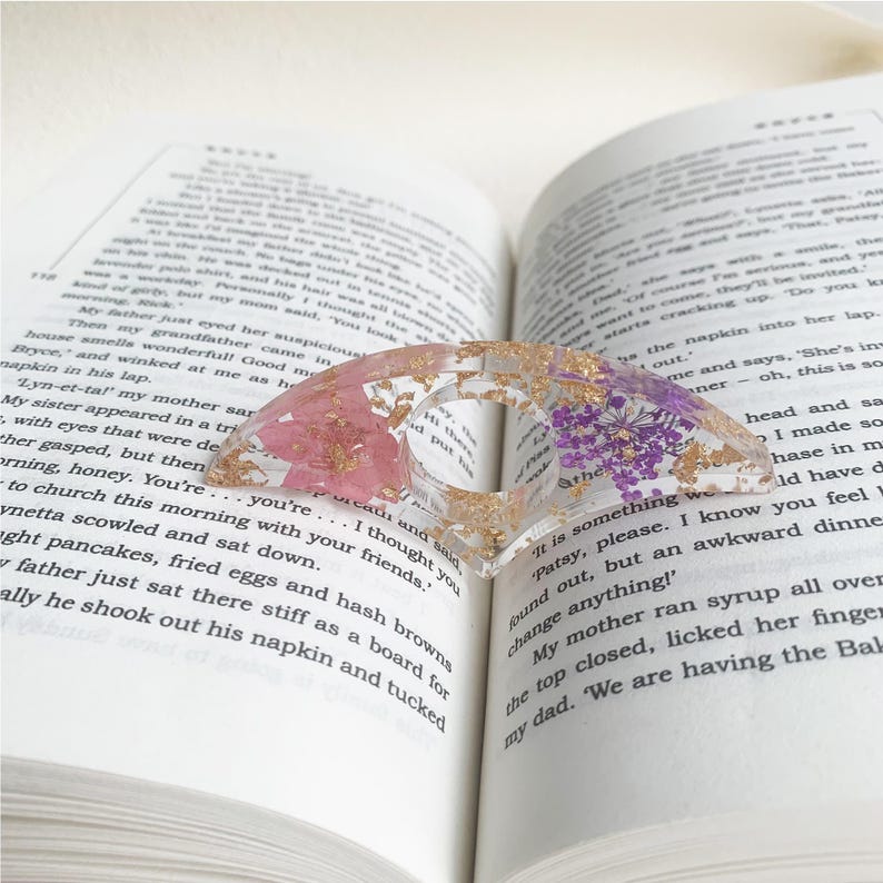 A resin book page holder featuring pink and purple flowers with gold flecks, placed between the pages of an open book. The holder delicately holds the pages in place, adding a beautiful and elegant touch to the reading experience