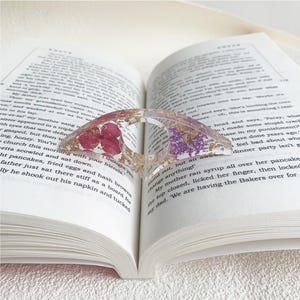 A resin book page holder featuring purple and pink flowers with gold flecks, embedded in clear resin, placed between the pages of an open book. The holder gently holds the pages in place, adding a delicate and vibrant touch to the reading experience