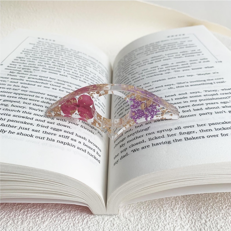 A resin book page holder featuring purple and pink flowers with gold flecks, embedded in clear resin, placed between the pages of an open book. The holder gently holds the pages in place, adding a delicate and vibrant touch to the reading experience