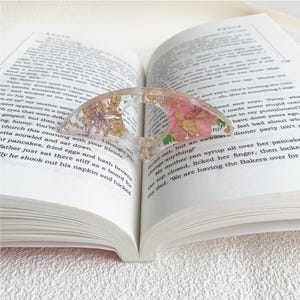 A resin book page holder featuring pink flowers and gold flecks, placed between the pages of an open book. The holder gently keeps the pages in place, adding a delicate and vibrant touch to the reading experience