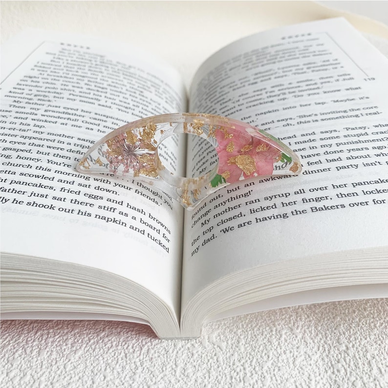 A resin book page holder featuring pink flowers and gold flecks, placed between the pages of an open book. The holder gently keeps the pages in place, adding a delicate and vibrant touch to the reading experience