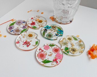 May include: Set of six clear resin car coasters with dried flowers and gold leaf accents. Each coaster features a different floral arrangement in shades of pink, purple, yellow, and blue. A textured glass is in the background.