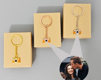 May include: Three keychains with miniature camera pendants. Each keychain has a different metal finish: gold, rose gold, and silver. The camera pendants are orange and black with a silver lens. They are displayed on small, beige boxes.