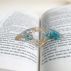 A resin book page holder featuring blue flowers and gold flecks, embedded in clear resin, placed between the pages of an open book. The holder gently keeps the pages in place, adding a calming and elegant touch to the reading experience