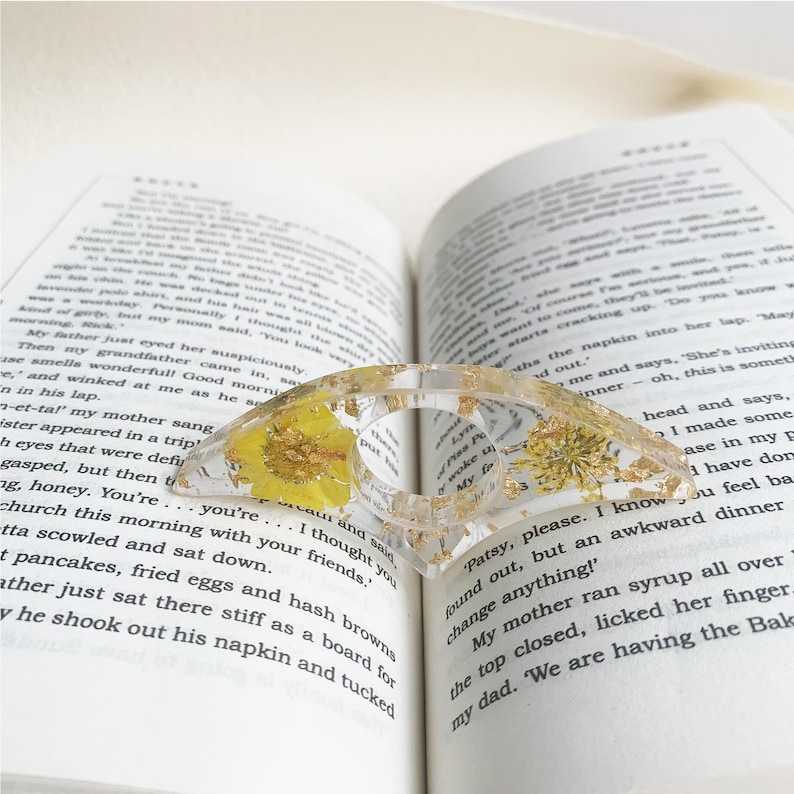 A resin book page holder featuring yellow flowers and gold flecks embedded in clear resin, placed between the pages of an open book. The holder gently keeps the pages in place, adding a bright and elegant touch to the reading experience