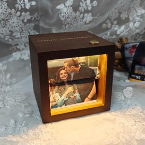 May include: A dark brown wooden photo cube with a lit interior, displaying a couple kissing. The top of the cube has the words "Happy Anniversary" engraved. A small gold button is on the top.