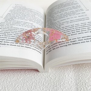 A resin book page holder featuring pink flowers and gold flecks embedded in clear resin, placed between the pages of an open book. The holder delicately keeps the pages in place, adding a soft and elegant touch to the reading experience