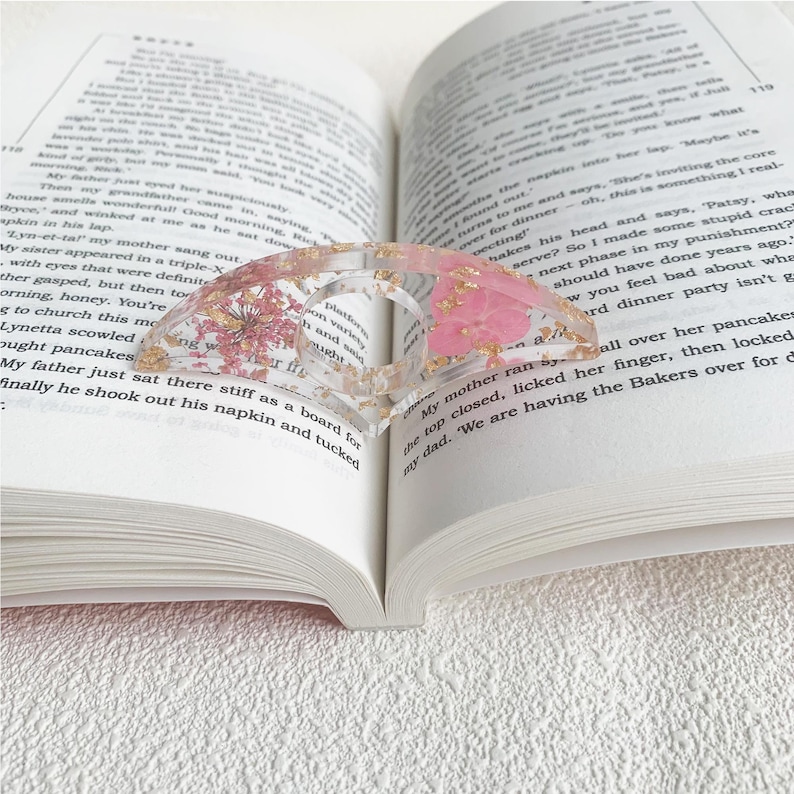 A resin book page holder featuring pink flowers and gold flecks embedded in clear resin, placed between the pages of an open book. The holder delicately keeps the pages in place, adding a soft and elegant touch to the reading experience