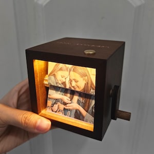 May include: A dark brown wooden photo cube with a lit-up image of a mother and child. The cube has a small crank on the side and a gold-colored button on top. The image is visible through a rectangular opening.