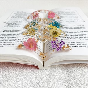 Four resin book page holders featuring different colored flowers (pink, blue, yellow, and purple) with gold flecks, placed between the pages of an open book. The holders gently keep the pages in place, adding a colorful and elegant touch to the reading experience