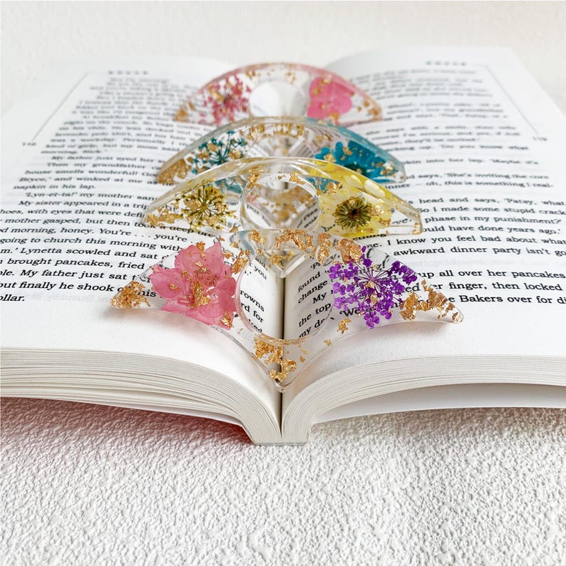 Four resin book page holders featuring different colored flowers (pink, blue, yellow, and purple) with gold flecks, placed between the pages of an open book. The holders gently keep the pages in place, adding a colorful and elegant touch to the reading experience