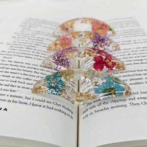 Op de afbeelding: Vier transparante hars boekenleggers met gedroogde bloemen en goudvlokken. Elke houder heeft een ander kleurenschema, waaronder roze, paarse, rode en blauwe bloemen. De houders liggen op de pagina's van een open boek.