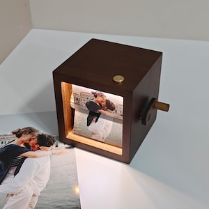 May include: A dark brown wooden photo cube with a lit-up image of a couple kissing. The cube has a small brass button on top and a wooden crank on the side. The cube is sitting on a white surface.