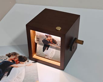 May include: A dark brown wooden photo cube with a lit-up image of a couple kissing. The cube has a small brass button on top and a wooden crank on the side. The cube is sitting on a white surface.