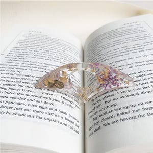 A resin book page holder featuring purple and gold flowers with flecks of gold, embedded in clear resin, placed between the pages of an open book. The holder gently keeps the pages in place, adding a refined and elegant touch to the reading experience
