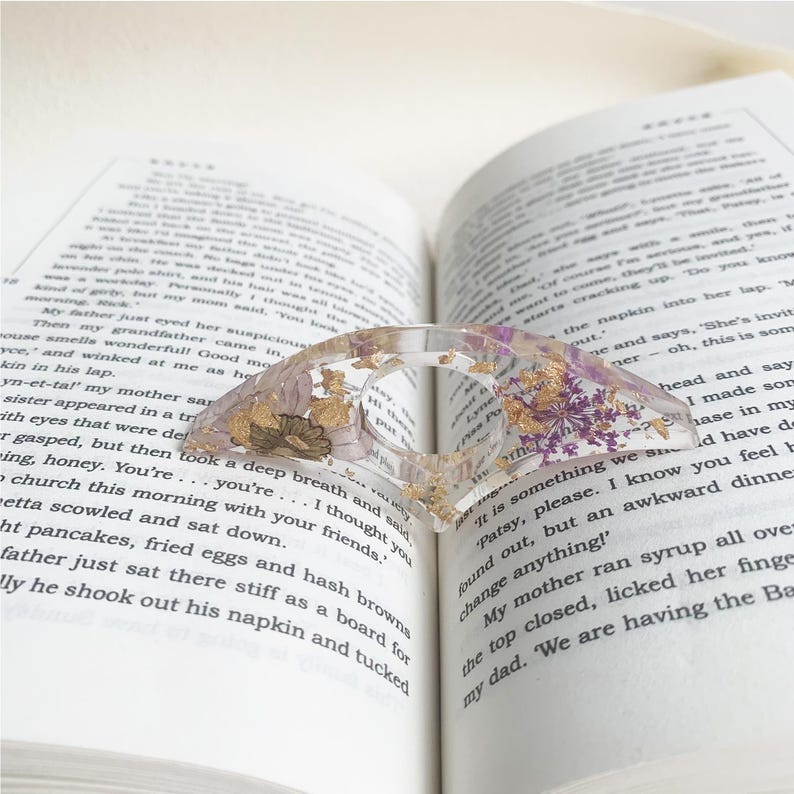 A resin book page holder featuring purple and gold flowers with flecks of gold, embedded in clear resin, placed between the pages of an open book. The holder gently keeps the pages in place, adding a refined and elegant touch to the reading experience