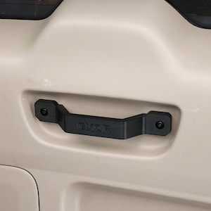 May include: A black handle with the text "GXOR" is mounted on a beige surface. The handle is rectangular with rounded edges and is secured with screws at each end. The background is a vehicle interior.