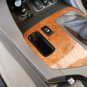 May include: Interior car console with wood grain trim. Features a black storage compartment, a button labeled "DAC", and a gear shift. The console is gray and brown, with a leather gear shift boot.