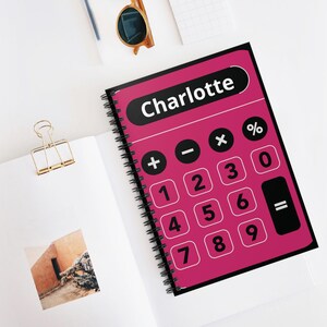 May include: A spiral-bound notebook featuring a calculator design. The cover is a vibrant pink with black calculator buttons and the name "Charlotte" in white. Other items on the white surface include sunglasses and a gold binder clip.
