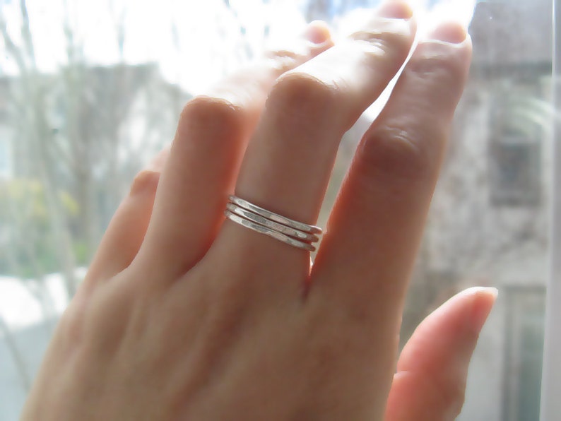 Puede incluir: Primer plano de una mano con tres anillos apilables de plata. Los anillos son finos y tienen una textura martillada.