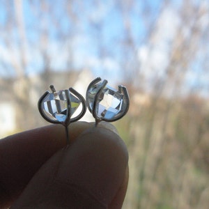 May include: A pair of stud earrings featuring clear, faceted, diamond-shaped stones set in silver-toned metal prongs. The stones are transparent and reflect light, held by a four-prong setting that secures the gem in place with a post backing.