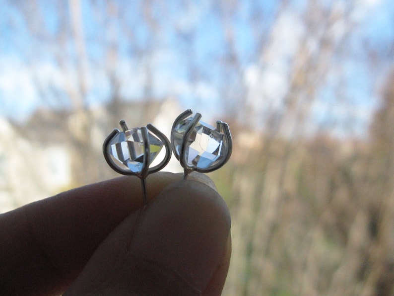 May include: A pair of stud earrings featuring clear, faceted, diamond-shaped stones set in silver-toned metal prongs. The stones are transparent and reflect light, held by a four-prong setting that secures the gem in place with a post backing.