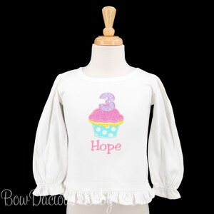 May include: White long-sleeved shirt with a pink cupcake with a purple number 3 on top. The cupcake has white polka dots on a blue background. The shirt has a ruffle at the bottom and the name "Hope" is embroidered below the cupcake.