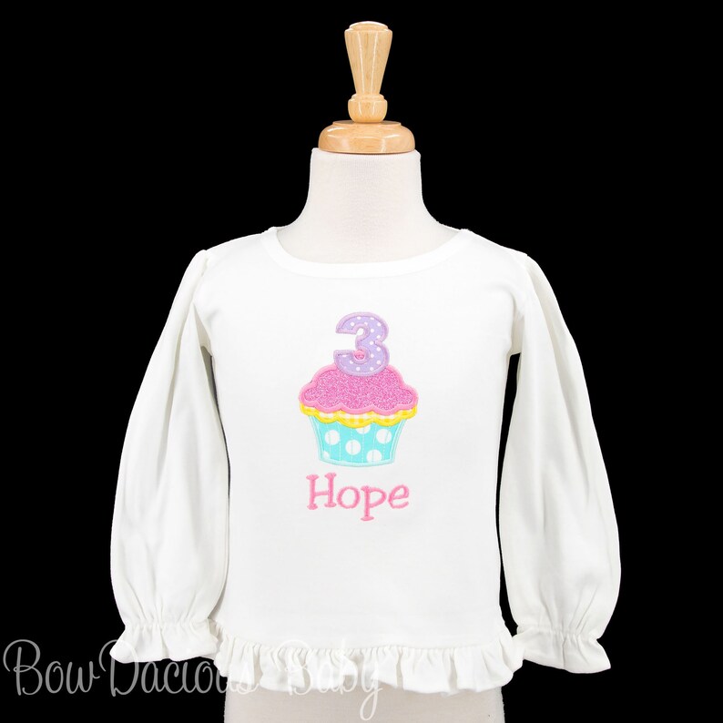 May include: White long-sleeved shirt with a pink cupcake with a purple number 3 on top. The cupcake has white polka dots on a blue background. The shirt has a ruffle at the bottom and the name "Hope" is embroidered below the cupcake.