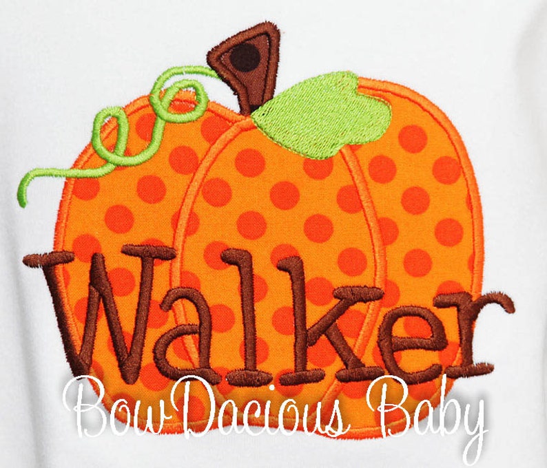 May include: Embroidered orange pumpkin with brown polka dots, green stem, and the name "Walker" in brown thread. The text "Bow Dacious Baby" is below the pumpkin. The design is on a white background.