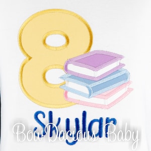 May include: A white shirt with a yellow number eight and a stack of colorful books. The text "Skylar Bow Dacious Baby" is embroidered in blue below the books.