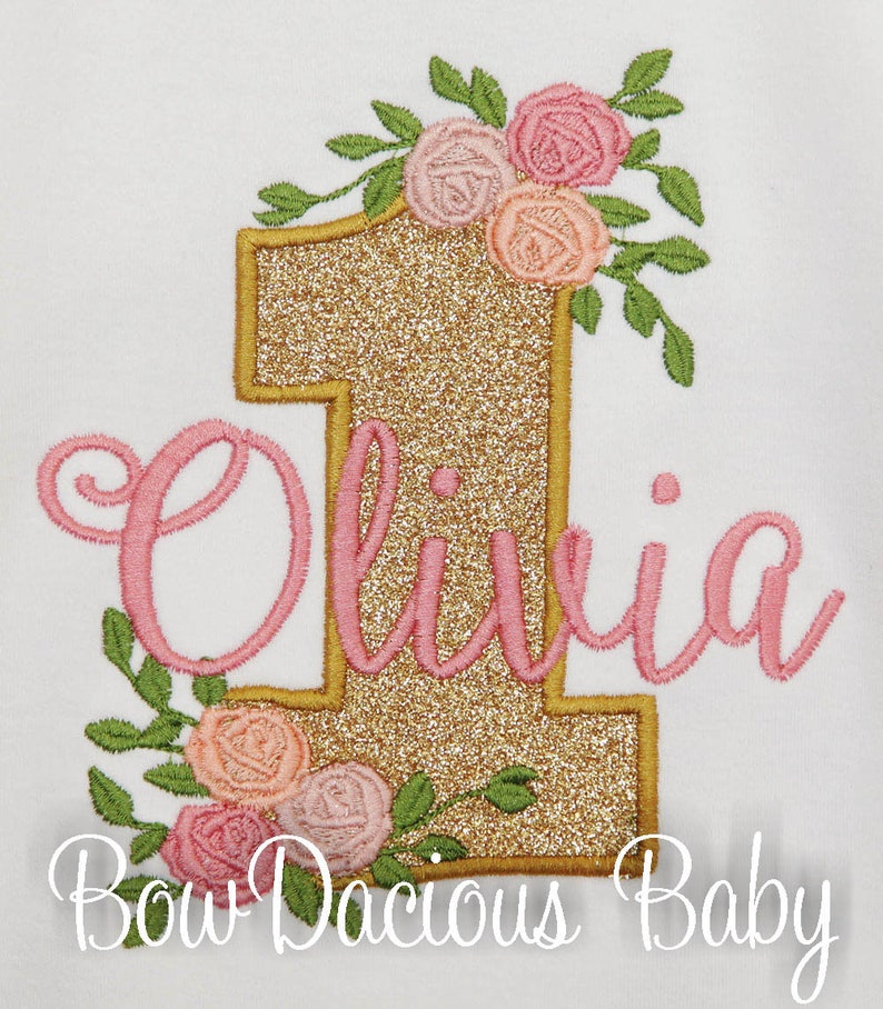 May include: Gold glitter number one with pink floral embellishments and the name Olivia in pink cursive script. The text "Bow Dacious Baby" is below the number.