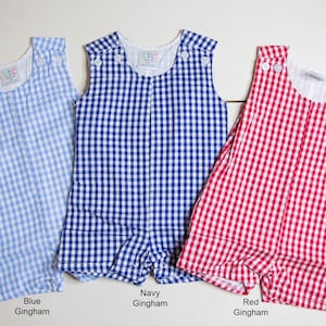 May include: Three sleeveless gingham rompers in blue, navy, and red. The rompers have a white background with a checkered pattern. The text "Blue Gingham", "Navy Gingham", and "Red Gingham" is printed below each romper.