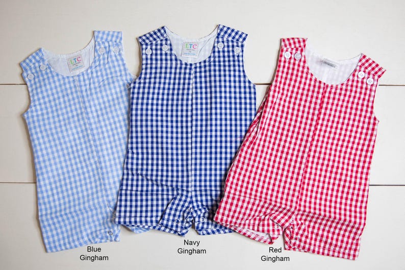 May include: Three sleeveless gingham rompers in blue, navy, and red. The rompers have a white background with a checkered pattern. The text "Blue Gingham", "Navy Gingham", and "Red Gingham" is printed below each romper.