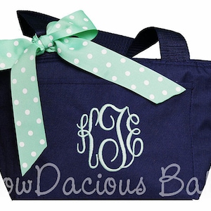 K&ouml;nnte beinhalten: Eine marineblaue Tragetasche mit einer gr&uuml;n-wei&szlig;en Polka-Dot-Schleife und einem monogrammierten Initial. Die Tasche hat einen Rei&szlig;verschluss und zwei Henkel.