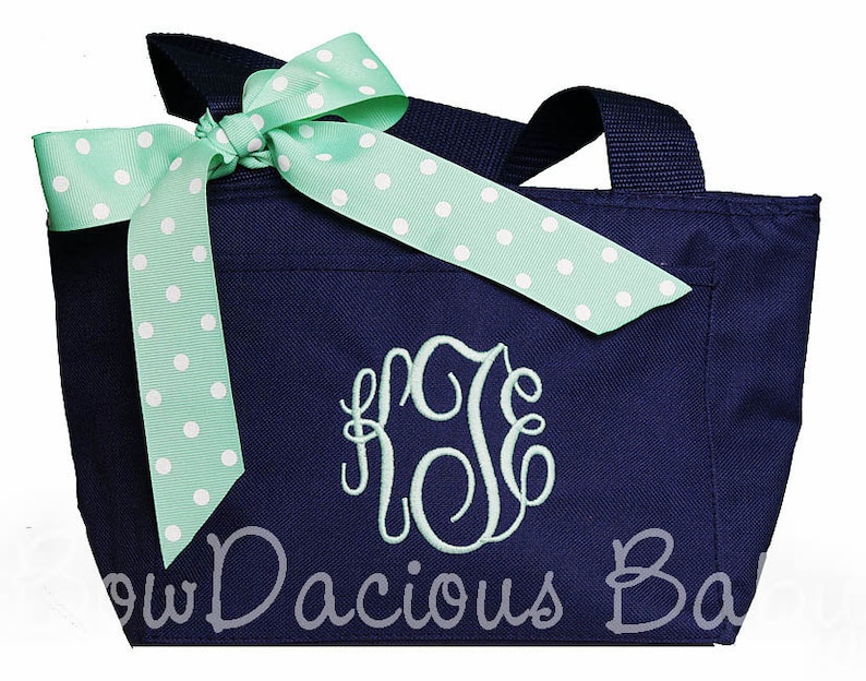 K&ouml;nnte beinhalten: Eine marineblaue Tragetasche mit einer gr&uuml;n-wei&szlig;en Polka-Dot-Schleife und einem monogrammierten Initial. Die Tasche hat einen Rei&szlig;verschluss und zwei Henkel.