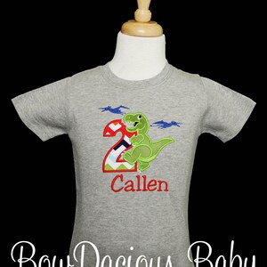 May include: A grey short-sleeved t-shirt with a green dinosaur and the number 2, with the name "Callen" in red. The shirt also has two blue pterodactyls. The text "Bow Dacious Baby" is at the bottom.