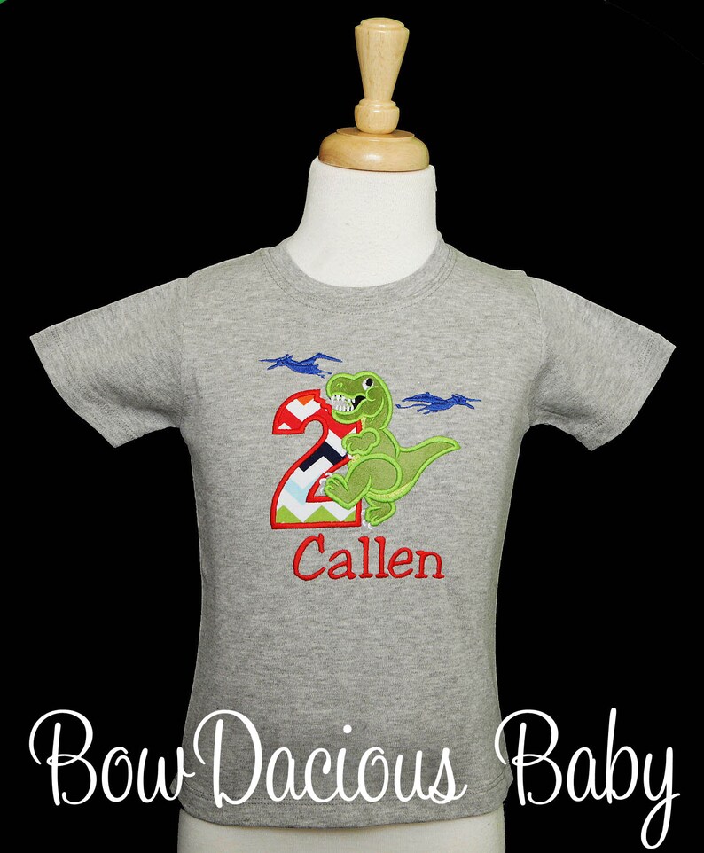 May include: A grey short-sleeved t-shirt with a green dinosaur and the number 2, with the name "Callen" in red. The shirt also has two blue pterodactyls. The text "Bow Dacious Baby" is at the bottom.