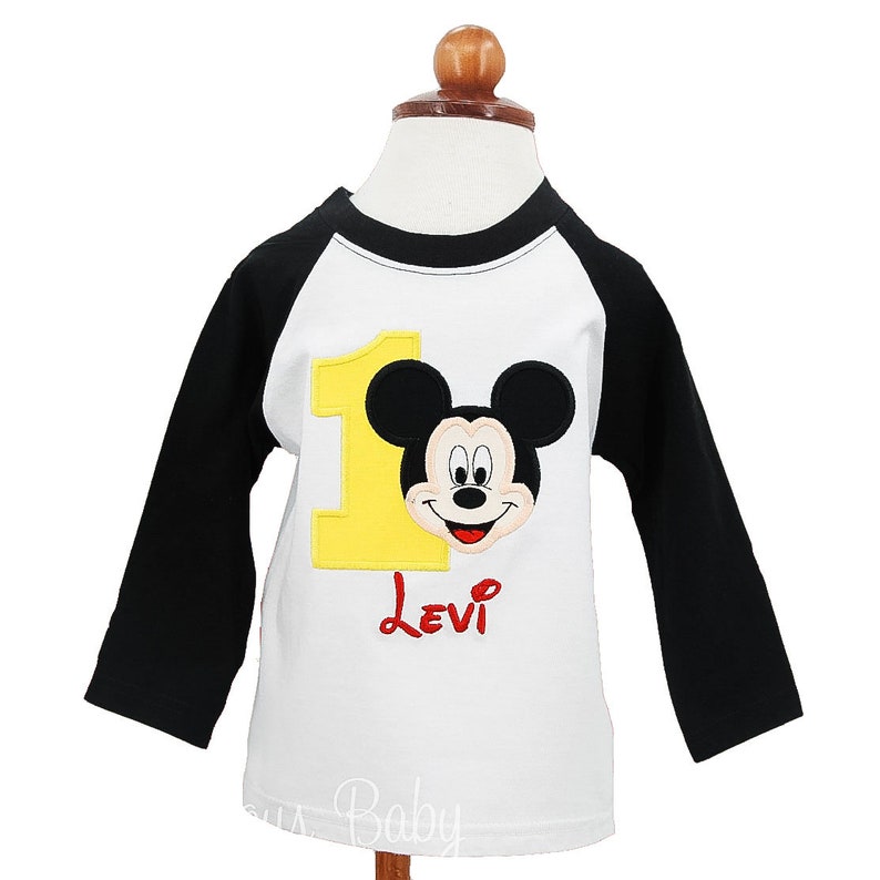 May include: White and black long-sleeved shirt featuring Mickey Mouse. The shirt has a yellow number 1 and the name "Levi" in red. The shirt is designed for a child's first birthday.