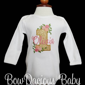 May include: White long-sleeve onesie with gold glitter number one and pink floral embroidery. The name "Olivia" is embroidered in pink below the number one.