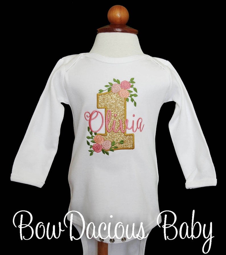 May include: White long-sleeve onesie with gold glitter number one and pink floral embroidery. The name "Olivia" is embroidered in pink below the number one.