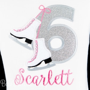 May include: A white shirt with a silver glitter number six and two white ice skates with pink accents. The name "Scarlett" is embroidered in pink below the skates.