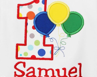 May include: A white onesie with a red outlined number one with colorful polka dots. Three balloons are attached to the number one, one yellow, one blue, and one green. The name Samuel is embroidered in red below the number one.