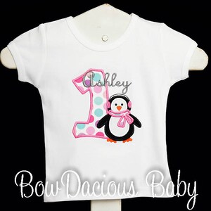 May include: White short-sleeved shirt with a pink and polka-dotted number 1 and the name "Ashley" in cursive. A penguin wearing earmuffs and a pink scarf is also featured. The text "Bow Dacious Baby" is at the bottom.
