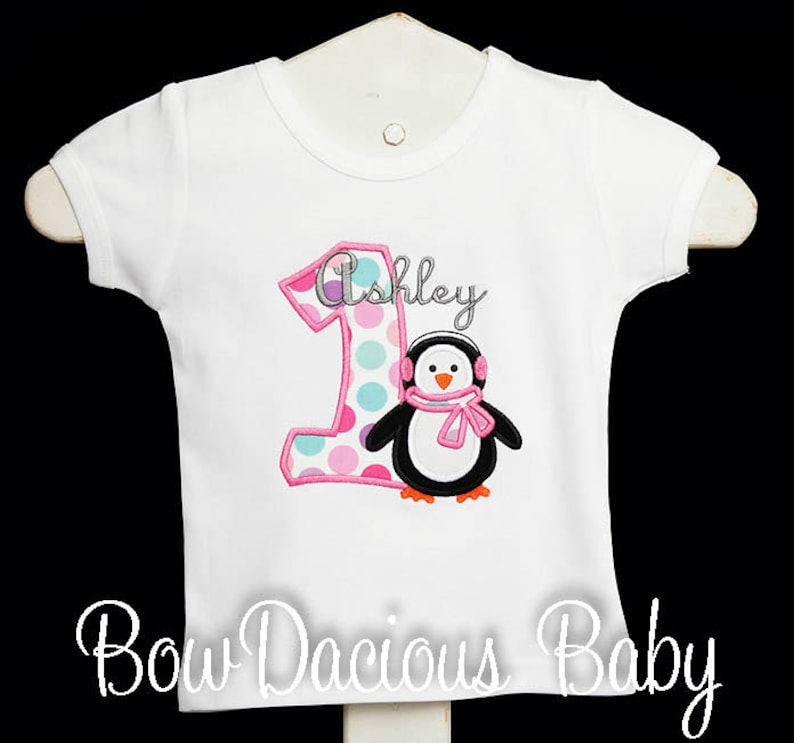 May include: White short-sleeved shirt with a pink and polka-dotted number 1 and the name "Ashley" in cursive. A penguin wearing earmuffs and a pink scarf is also featured. The text "Bow Dacious Baby" is at the bottom.