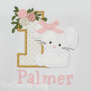 May include: A gold number one with white polka dots and a white bunny with a pink bow. The bunny is sitting next to the number one. The name Palmer is embroidered in pink below the number one.