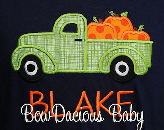May include: A navy blue shirt featuring an embroidered green truck carrying orange pumpkins with polka dots. The name "BLAKE" is embroidered in orange, with "Bow Dacious Baby" in white script below.