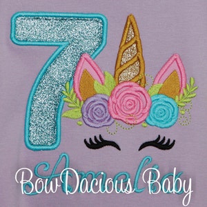 Puede incluir: Una camiseta morada con un n&uacute;mero 7 turquesa y una cabeza de unicornio con un cuerno dorado y una corona de flores. El texto "Bow Dacious Baby" est&aacute; debajo del unicornio.
