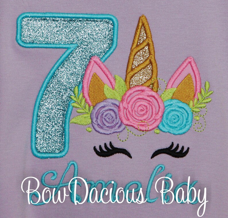 Puede incluir: Una camiseta morada con un n&uacute;mero 7 turquesa y una cabeza de unicornio con un cuerno dorado y una corona de flores. El texto "Bow Dacious Baby" est&aacute; debajo del unicornio.