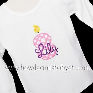 May include: White long-sleeved shirt with a pink number eight with white polka dots and a yellow flame on top. The number eight has the name "Lily" written in purple cursive.